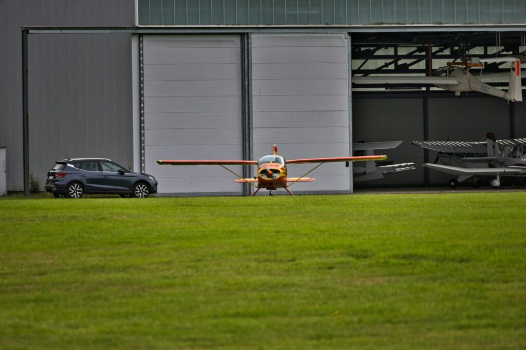 Small aircraft positioned on a grass runway beside a modern car and open hangar.
