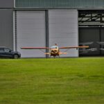 Small aircraft positioned on a grass runway beside a modern car and open hangar.